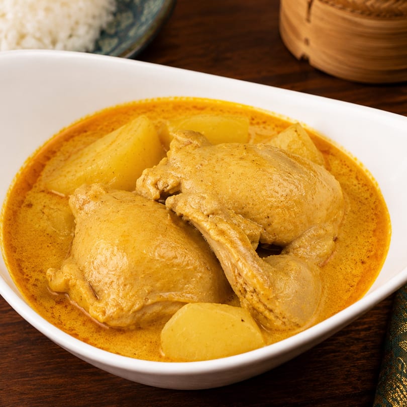 Chicken Massaman curry with potatoes in a white bowl, with rice and a bamboo steamer in the background.